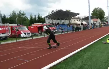 Grupa strażak&oacute;w w czarnych mundurach z odblaskowymi elementami stoi w zwartym szyku na zielonym boisku sportowym. W tle widać wysokie maszty oświetleniowe oraz drzewa i niebo z chmurami. Kilka os&oacute;b w środku grupy ubranych jest na czarno, ale bez odblask&oacute;w