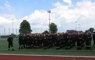 Grupa strażak&oacute;w w czarnych mundurach z odblaskowymi elementami stoi w zwartym szyku na zielonym boisku sportowym. W tle widać wysokie maszty oświetleniowe oraz drzewa i niebo z chmurami. Kilka os&oacute;b w środku grupy ubranych jest na czarno, ale bez odblask&oacute;w
