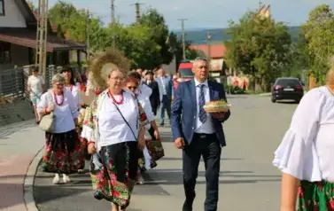 Poch&oacute;d dożynkowy. Korow&oacute;d wielu os&oacute;b ubranych elegancko, a także w stroje ludowe. Kobiety niosą dożynkowe wieńce. Niosą także chleby