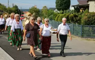 Poch&oacute;d dożynkowy. Korow&oacute;d wielu os&oacute;b ubranych elegancko, a także w stroje ludowe. Kobiety niosą dożynkowe wieńce. Niosą także chleby