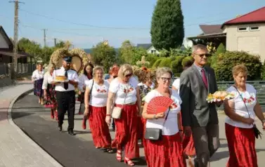 Poch&oacute;d dożynkowy. Korow&oacute;d wielu os&oacute;b ubranych elegancko, a także w stroje ludowe. Kobiety niosą dożynkowe wieńce. Niosą także chleby