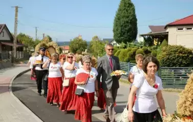 Poch&oacute;d dożynkowy. Korow&oacute;d wielu os&oacute;b ubranych elegancko, a także w stroje ludowe. Kobiety niosą dożynkowe wieńce. Niosą także chleby
