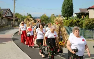 Poch&oacute;d dożynkowy. Korow&oacute;d wielu os&oacute;b ubranych elegancko, a także w stroje ludowe. Kobiety niosą dożynkowe wieńce. Niosą także chleby