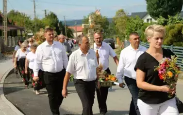 Poch&oacute;d dożynkowy. Korow&oacute;d wielu os&oacute;b ubranych elegancko, a także w stroje ludowe. Kobiety niosą dożynkowe wieńce. Niosą także chleby