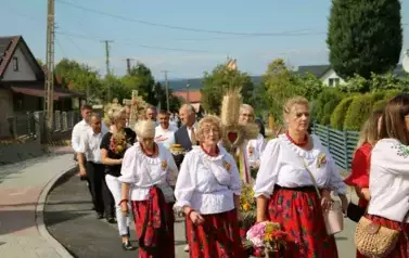 Poch&oacute;d dożynkowy. Korow&oacute;d wielu os&oacute;b ubranych elegancko, a także w stroje ludowe. Kobiety niosą dożynkowe wieńce. Niosą także chleby