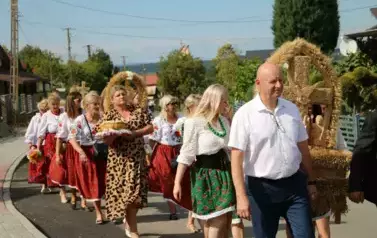 Poch&oacute;d dożynkowy. Korow&oacute;d wielu os&oacute;b ubranych elegancko, a także w stroje ludowe. Kobiety niosą dożynkowe wieńce. Niosą także chleby