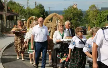 Poch&oacute;d dożynkowy. Korow&oacute;d wielu os&oacute;b ubranych elegancko, a także w stroje ludowe. Kobiety niosą dożynkowe wieńce. Niosą także chleby