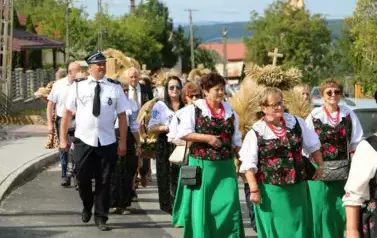 Poch&oacute;d dożynkowy. Korow&oacute;d wielu os&oacute;b ubranych elegancko, a także w stroje ludowe. Kobiety niosą dożynkowe wieńce. Niosą także chleby