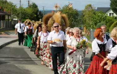 Poch&oacute;d dożynkowy. Korow&oacute;d wielu os&oacute;b ubranych elegancko, a także w stroje ludowe. Kobiety niosą dożynkowe wieńce. Niosą także chleby