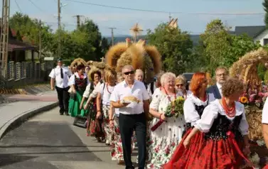 Poch&oacute;d dożynkowy. Korow&oacute;d wielu os&oacute;b ubranych elegancko, a także w stroje ludowe. Kobiety niosą dożynkowe wieńce. Niosą także chleby