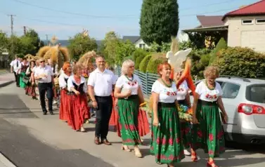 Poch&oacute;d dożynkowy. Korow&oacute;d wielu os&oacute;b ubranych elegancko, a także w stroje ludowe. Kobiety niosą dożynkowe wieńce. Niosą także chleby