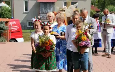 Poch&oacute;d dożynkowy. Korow&oacute;d wielu os&oacute;b ubranych elegancko, a także w stroje ludowe. Kobiety niosą dożynkowe wieńce. Niosą także chleby