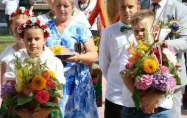 Poch&oacute;d dożynkowy. Korow&oacute;d wielu os&oacute;b ubranych elegancko, a także w stroje ludowe. Kobiety niosą dożynkowe wieńce. Niosą także chleby