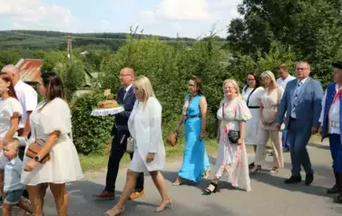 Poch&oacute;d dożynkowy. Korow&oacute;d wielu os&oacute;b ubranych elegancko, a także w stroje ludowe. Kobiety niosą dożynkowe wieńce. Niosą także chleby