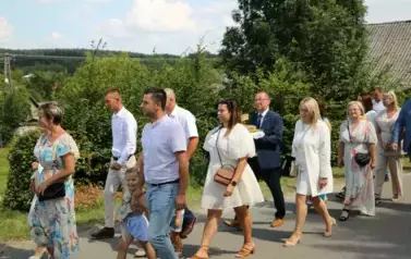 Poch&oacute;d dożynkowy. Korow&oacute;d wielu os&oacute;b ubranych elegancko, a także w stroje ludowe. Kobiety niosą dożynkowe wieńce. Niosą także chleby