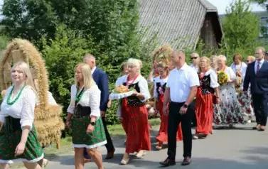 Poch&oacute;d dożynkowy. Korow&oacute;d wielu os&oacute;b ubranych elegancko, a także w stroje ludowe. Kobiety niosą dożynkowe wieńce. Niosą także chleby