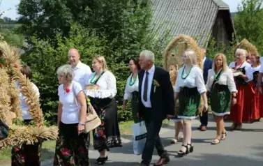 Poch&oacute;d dożynkowy. Korow&oacute;d wielu os&oacute;b ubranych elegancko, a także w stroje ludowe. Kobiety niosą dożynkowe wieńce. Niosą także chleby