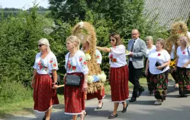 Poch&oacute;d dożynkowy. Korow&oacute;d wielu os&oacute;b ubranych elegancko, a także w stroje ludowe. Kobiety niosą dożynkowe wieńce. Niosą także chleby