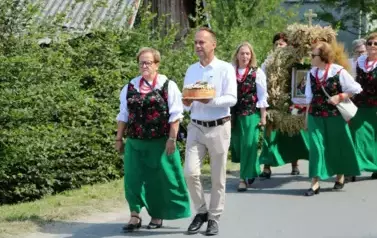 Poch&oacute;d dożynkowy. Korow&oacute;d wielu os&oacute;b ubranych elegancko, a także w stroje ludowe. Kobiety niosą dożynkowe wieńce. Niosą także chleby