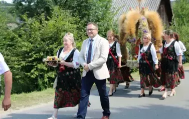 Poch&oacute;d dożynkowy. Korow&oacute;d wielu os&oacute;b ubranych elegancko, a także w stroje ludowe. Kobiety niosą dożynkowe wieńce. Niosą także chleby