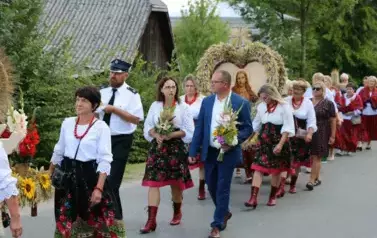Poch&oacute;d dożynkowy. Korow&oacute;d wielu os&oacute;b ubranych elegancko, a także w stroje ludowe. Kobiety niosą dożynkowe wieńce. Niosą także chleby