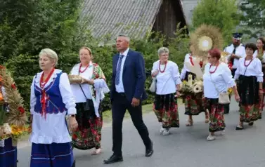 Poch&oacute;d dożynkowy. Korow&oacute;d wielu os&oacute;b ubranych elegancko, a także w stroje ludowe. Kobiety niosą dożynkowe wieńce. Niosą także chleby