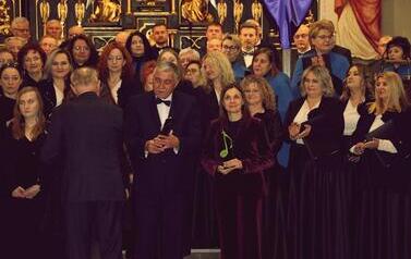 Portretowe ujęcie dyrygentki Magdaleny Makiel, ubranej w elegancką, welurową fioletową marynarkę. Kobieta patrzy w stronę obiektywu, uśmiechając się delikatnie. W obu dłoniach trzyma pamiątkową, zieloną statuetkę w kształcie klucza wiolinowego, kt&oacute;ra stoi