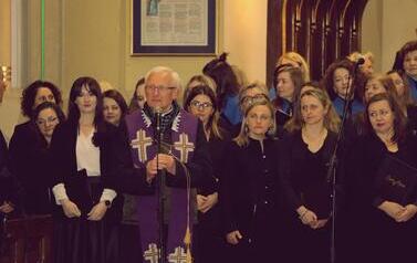 Zbliżenie na grupę os&oacute;b po koncercie. Po lewej stoi starszy kapłan w fioletowym ornacie, ściskający dłoń dyrygentki Magdaleny Makiel, kt&oacute;ra ubrana jest w elegancką fioletową marynarkę. Dyrygentka drugą ręką trzyma pamiątkową, zieloną statuetkę w kształcie