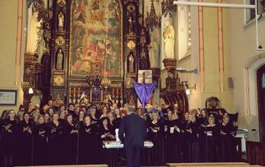 Zdjęcie przedstawia bardzo liczną grupę ch&oacute;rzyst&oacute;w wypełniającą całe prezbiterium kościoła, śpiewających wsp&oacute;lnie. Są ustawieni w wielu rzędach przed tym samym bogatym ołtarzem, co na poprzednim zdjęciu. Dyrygent, mężczyzna w ciemnym garniturze, stoi tyłe