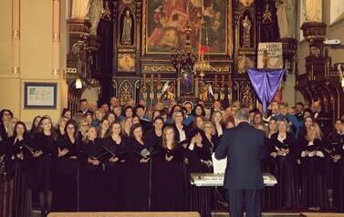 Zdjęcie przedstawia bardzo liczną grupę ch&oacute;rzyst&oacute;w wypełniającą całe prezbiterium kościoła, śpiewających wsp&oacute;lnie. Są ustawieni w wielu rzędach przed tym samym bogatym ołtarzem, co na poprzednim zdjęciu. Dyrygent, mężczyzna w ciemnym garniturze, stoi tyłe