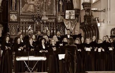 Szerokie ujęcie wnętrza zabytkowego kościoła w Korczynie, oświetlonego ciepłym światłem. Na stopniach przed monumentalnym, rzeźbionym, ciemnym ołtarzem stoi Ch&oacute;r Legato. Ch&oacute;rzyści (kobiety w długich czarnych sukniach, mężczyźni w garniturach) są ustawieni