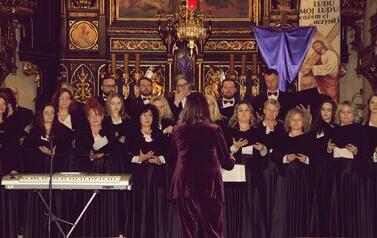 Szerokie ujęcie wnętrza zabytkowego kościoła w Korczynie, oświetlonego ciepłym światłem. Na stopniach przed monumentalnym, rzeźbionym, ciemnym ołtarzem stoi Ch&oacute;r Legato. Ch&oacute;rzyści (kobiety w długich czarnych sukniach, mężczyźni w garniturach) są ustawieni