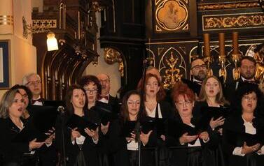 Szerokie ujęcie wnętrza zabytkowego kościoła w Korczynie, oświetlonego ciepłym światłem. Na stopniach przed monumentalnym, rzeźbionym, ciemnym ołtarzem stoi Ch&oacute;r Legato. Ch&oacute;rzyści (kobiety w długich czarnych sukniach, mężczyźni w garniturach) są ustawieni