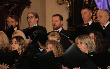 Szerokie ujęcie wnętrza zabytkowego kościoła w Korczynie, oświetlonego ciepłym światłem. Na stopniach przed monumentalnym, rzeźbionym, ciemnym ołtarzem stoi Ch&oacute;r Legato. Ch&oacute;rzyści (kobiety w długich czarnych sukniach, mężczyźni w garniturach) są ustawieni