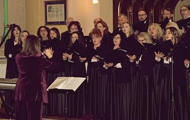 Szerokie ujęcie wnętrza zabytkowego kościoła w Korczynie, oświetlonego ciepłym światłem. Na stopniach przed monumentalnym, rzeźbionym, ciemnym ołtarzem stoi Ch&oacute;r Legato. Ch&oacute;rzyści (kobiety w długich czarnych sukniach, mężczyźni w garniturach) są ustawieni