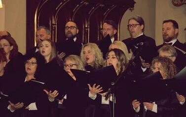 Szerokie ujęcie wnętrza zabytkowego kościoła w Korczynie, oświetlonego ciepłym światłem. Na stopniach przed monumentalnym, rzeźbionym, ciemnym ołtarzem stoi Ch&oacute;r Legato. Ch&oacute;rzyści (kobiety w długich czarnych sukniach, mężczyźni w garniturach) są ustawieni