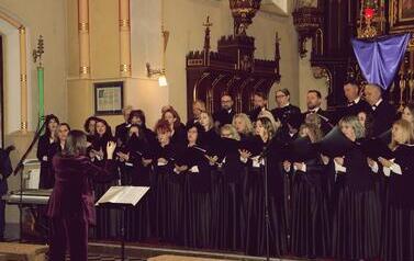 Szerokie ujęcie wnętrza zabytkowego kościoła w Korczynie, oświetlonego ciepłym światłem. Na stopniach przed monumentalnym, rzeźbionym, ciemnym ołtarzem stoi Ch&oacute;r Legato. Ch&oacute;rzyści (kobiety w długich czarnych sukniach, mężczyźni w garniturach) są ustawieni