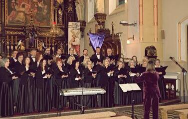 Szerokie ujęcie wnętrza zabytkowego kościoła w Korczynie, oświetlonego ciepłym światłem. Na stopniach przed monumentalnym, rzeźbionym, ciemnym ołtarzem stoi Ch&oacute;r Legato. Ch&oacute;rzyści (kobiety w długich czarnych sukniach, mężczyźni w garniturach) są ustawieni