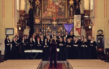 djęcie przedstawia bardzo liczną grupę ch&oacute;rzyst&oacute;w wypełniającą całe prezbiterium kościoła, śpiewających wsp&oacute;lnie. Są ustawieni w wielu rzędach przed tym samym bogatym ołtarzem, co na poprzednim zdjęciu. Dyrygent, mężczyzna w ciemnym garniturze, stoi tyłem