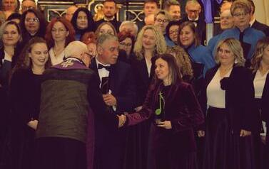 Zbliżenie na grupę os&oacute;b po koncercie. Po lewej stoi starszy kapłan w fioletowym ornacie, ściskający dłoń dyrygentki Magdaleny Makiel, kt&oacute;ra ubrana jest w elegancką fioletową marynarkę. Dyrygentka drugą ręką trzyma pamiątkową, zieloną statuetkę w kształcie