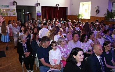 Publiczność zgromadzona w sali widowiskowej podczas lokalnego wydarzenia kulturalnego. Widzowie w r&oacute;żnym wieku siedzą na krzesłach, uważnie oglądając program artystyczny. W pierwszym rzędzie widoczne są zaproszone pary oraz przedstawiciele lokalnych władz