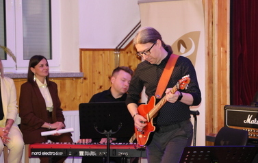 Muzycy akompaniujący artystom podczas koncertu. Na pierwszym planie widać perkusistę oraz basistę grających na swoich instrumentach. W tle, na podwyższeniu, widoczni są członkowie ch&oacute;ru oraz kwiatowe dekoracje sceny.