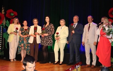 Grupa wyr&oacute;żnionych kobiet stojących w rzędzie na scenie. Każda z nich trzyma duży, bogaty bukiet kwiat&oacute;w oraz pamiątkowy folder. Obok nich stoją organizatorzy i przedstawiciele władz podsumowujący tę część uroczystości.