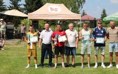 Seria dynamicznych i pozowanych zdjęć z amatorskiego turnieju piłki nożnej, kt&oacute;ry odbył się w pogodny, słoneczny dzień na trawiastym boisku otoczonym brzozami. Zdjęcia przedstawiają pełny przebieg wydarzenia sportowego. Galeria zawiera pozowane fotografie