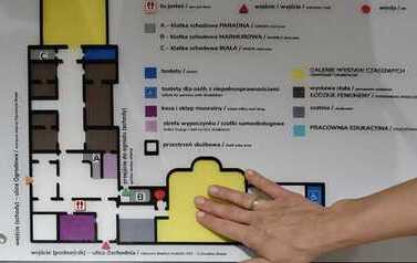 Plan orientacyjny Pałacu Poznańskiego w Łodzi (Muzeum Miasta Łodzi). Plan jest wykonany w formie tyflograficznej z wyczuwalnymi fakturami i wyraźnymi kolorami (np. czerwony punkt &bdquo;tu jesteś&rdquo;, zielona &bdquo;strefa wyciszenia&rdquo;). Poniżej znajdują się piktogramy z