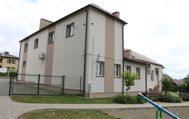 Zestaw 6 zdjęć przedstawia odnowioną elewację budynku Domu Ludowego w Zręcinie po zakończeniu prac remontowych. Budynek posiada nowoczesne wykończenie w jasnej, kremowo-beżowej kolorystyce z ciemniejszymi, beżowo-brązowymi pasami akcentującymi oraz brązow