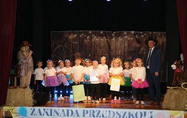 Zbliżenie na grupę dzieci stojących na scenie przed tłem przedstawiającym drzewa i światełka. Dzieci są ubrane w białe koszulki i sp&oacute;dniczki tiulowe w r&oacute;żnych jaskrawych kolorach (fioletowy, niebieski, ż&oacute;łty, r&oacute;żowy, zielony). Stoją przed mikrofonami, zapewne śpiewając.