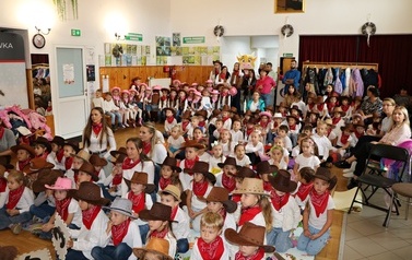 Zdjęcie z szerokiego planu przedstawia dużą grupę dzieci, prawdopodobnie w wieku przedszkolnym, siedzących na podłodze w sali. Większość ma na sobie białe koszulki, czerwone chustki na szyjach i brązowe kapelusze kowbojskie. Z tyłu widać dorosłych opiekun&oacute;w oraz dekoracje sali z plakatami i oknami.