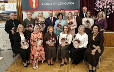 Grupa kilkunastu dyrektor&oacute;w i przedstawicieli plac&oacute;wek oświatowych pozuje do pamiątkowej fotografii na tle ścianki promocyjnej &bdquo;Gmina Chork&oacute;wka&rdquo;. Osoby z pierwszego rzędu siedzą na krzesłach, a za nimi stoi drugi rząd uczestnik&oacute;w spotkania. Prawie wszyscy obecni trzymają w rękach dekoracyjne kompozycje z ozdobnych wrzos&oacute;w w doniczkach zawiniętych w papier. W tle po prawej stronie widoczne jest ozdobne drzewko z r&oacute;żowymi kwiatami.