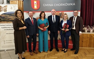 Sześć os&oacute;b na tle ścianki promocyjnej Gminy Chork&oacute;wka. Dwie wyr&oacute;żnione osoby w eleganckich strojach trzymają w rękach czerwone pamiątkowe dyplomy i r&oacute;że. Po bokach stoją przedstawiciele gminy oraz gospodarze uroczystości.